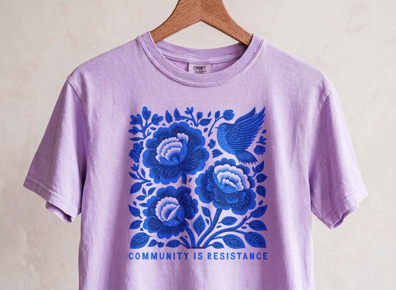 Puede incluir: Una camiseta de manga corta de color morado claro con un gr&aacute;fico cuadrado de flores azules y un p&aacute;jaro. Debajo de la ilustraci&oacute;n, el texto COMMUNITY IS RESISTANCE aparece en letras may&uacute;sculas azules. La camiseta cuelga de una percha de madera.