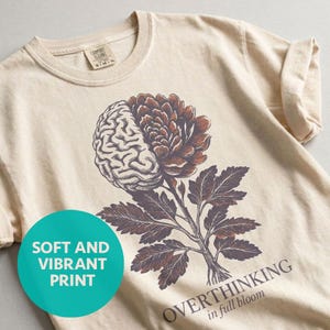 May include: Cream-colored t-shirt featuring a graphic of a brain and flower. The brain is on the left, and the flower is on the right. The text "OVERTHINKING in full bloom" is printed at the bottom. A teal circle with the words "SOFT AND VIBRANT PRINT" is on the left.