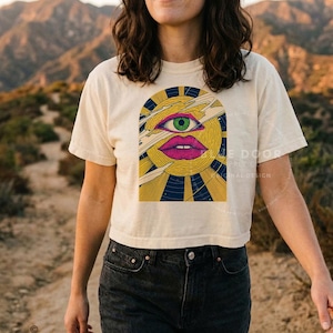 Woman wearing a witchy boxy crop tee in ivory, psychedelic sun with a green third eye, engraved comets and navy star field. She's walking in the desert.