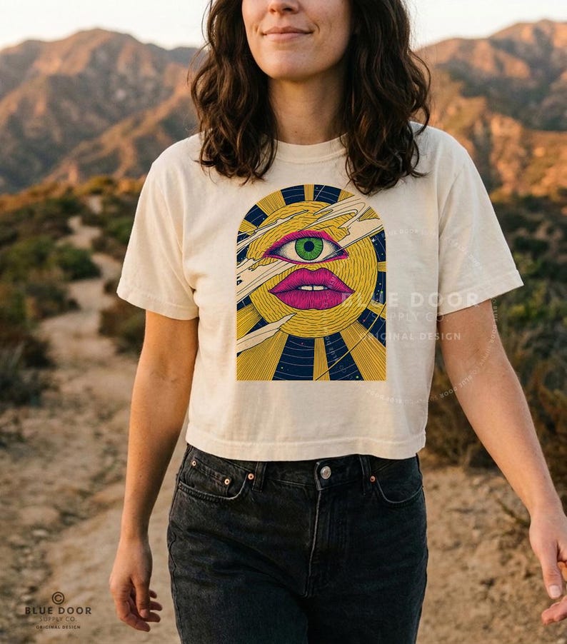 Woman wearing a witchy boxy crop tee in ivory, psychedelic sun with a green third eye, engraved comets and navy star field. She's walking in the desert.