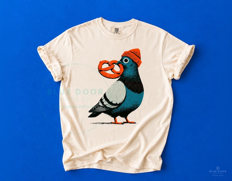 NYC shirt in ivory on a bold cobalt background, funny city pigeon clutching an oversized soft pretzel