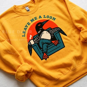 Loon crewneck sweatshirt in gold laid flat, funny loon gift featuring a bird relaxing in an armchair with coffee mug, Minnesota loon apparel for lake house weekends