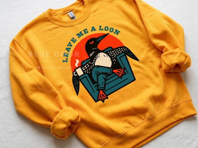 Loon crewneck sweatshirt in gold laid flat, funny loon gift featuring a bird relaxing in an armchair with coffee mug, Minnesota loon apparel for lake house weekends