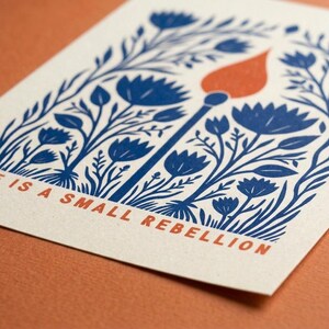 May include: A rectangular off-white card featuring a blue ink illustration of stylized flowers and foliage surrounding a single lit match with an orange flame. The text "HOPE IS A SMALL REBELLION" is printed in orange at the bottom of the card.