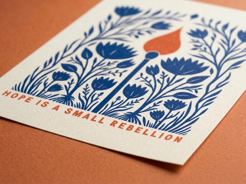 May include: A rectangular off-white card featuring a blue ink illustration of stylized flowers and foliage surrounding a single lit match with an orange flame. The text "HOPE IS A SMALL REBELLION" is printed in orange at the bottom of the card.