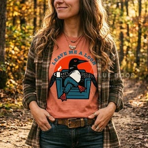 May include: A rust-colored t-shirt featuring a graphic of a loon sitting in a blue armchair, holding a white mug, with the text "LEAVE ME A LOON" arched above. The loon has black feathers, a white chest, and orange feet, set against a large orange circle.