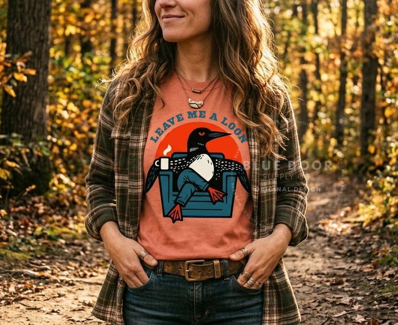 May include: A rust-colored t-shirt featuring a graphic of a loon sitting in a blue armchair, holding a white mug, with the text "LEAVE ME A LOON" arched above. The loon has black feathers, a white chest, and orange feet, set against a large orange circle.