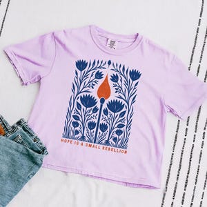 Orchid lavender Comfort Colors boxy crop tee laid flat with high-waisted jeans, featuring a folk art botanical illustration of a lit match with red-orange flame and the phrase Hope Is A Small Rebellion — liberal feminist graphic crop top.