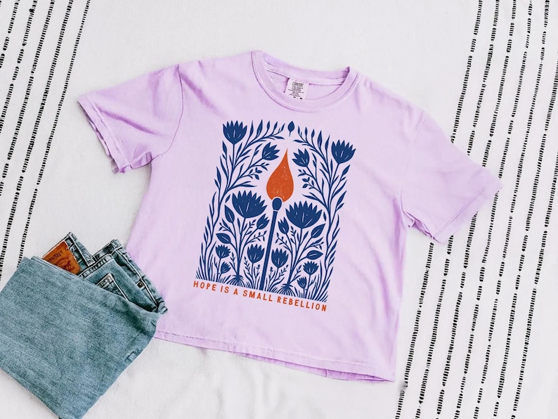 Orchid lavender Comfort Colors boxy crop tee laid flat with high-waisted jeans, featuring a folk art botanical illustration of a lit match with red-orange flame and the phrase Hope Is A Small Rebellion — liberal feminist graphic crop top.