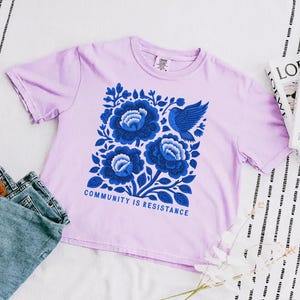 Puede incluir: Una camiseta de manga corta de color morado claro con un gr&aacute;fico central de tres flores grandes azules y un p&aacute;jaro azul. Debajo del dise&ntilde;o floral, el texto "COMMUNITY IS RESISTANCE" aparece en letras may&uacute;sculas azules. Cuello redondo.