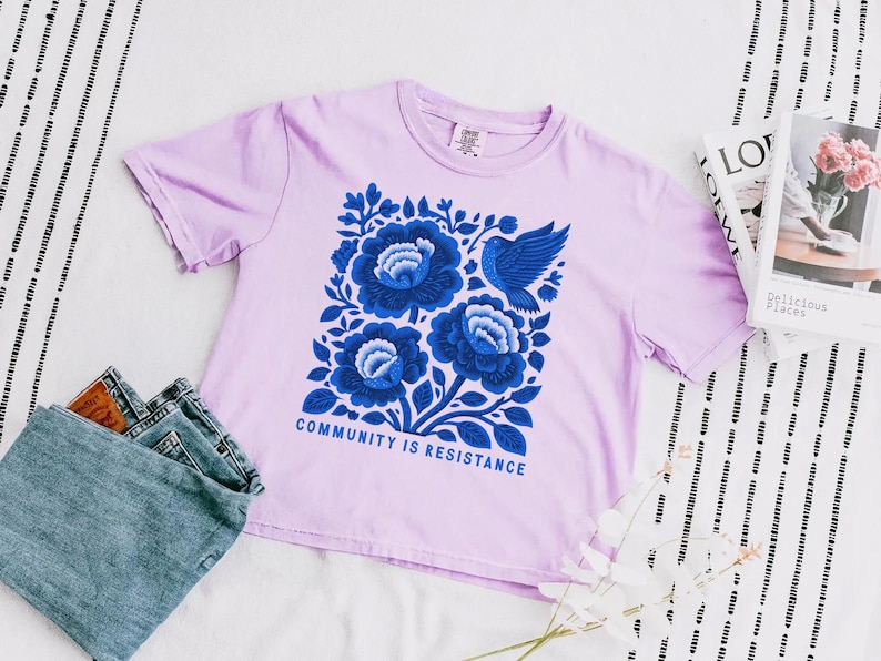 Puede incluir: Una camiseta de manga corta de color morado claro con un gr&aacute;fico central de tres flores grandes azules y un p&aacute;jaro azul. Debajo del dise&ntilde;o floral, el texto "COMMUNITY IS RESISTANCE" aparece en letras may&uacute;sculas azules. Cuello redondo.