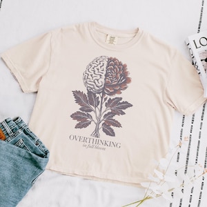 May include: Cream-colored t-shirt featuring a graphic of a brain and flower, with the text "OVERTHINKING in full bloom". The shirt is paired with blue jeans. A magazine is visible in the background.