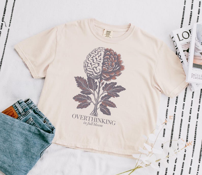 May include: Cream-colored t-shirt featuring a graphic of a brain and flower, with the text "OVERTHINKING in full bloom". The shirt is paired with blue jeans. A magazine is visible in the background.
