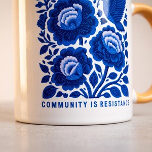 May include: A white ceramic mug with a yellow handle featuring a blue floral pattern of large blossoms and a bird. The text "COMMUNITY IS RESISTANCE" is printed in blue at the bottom of the design. The mug has a smooth, glossy finish throughout.