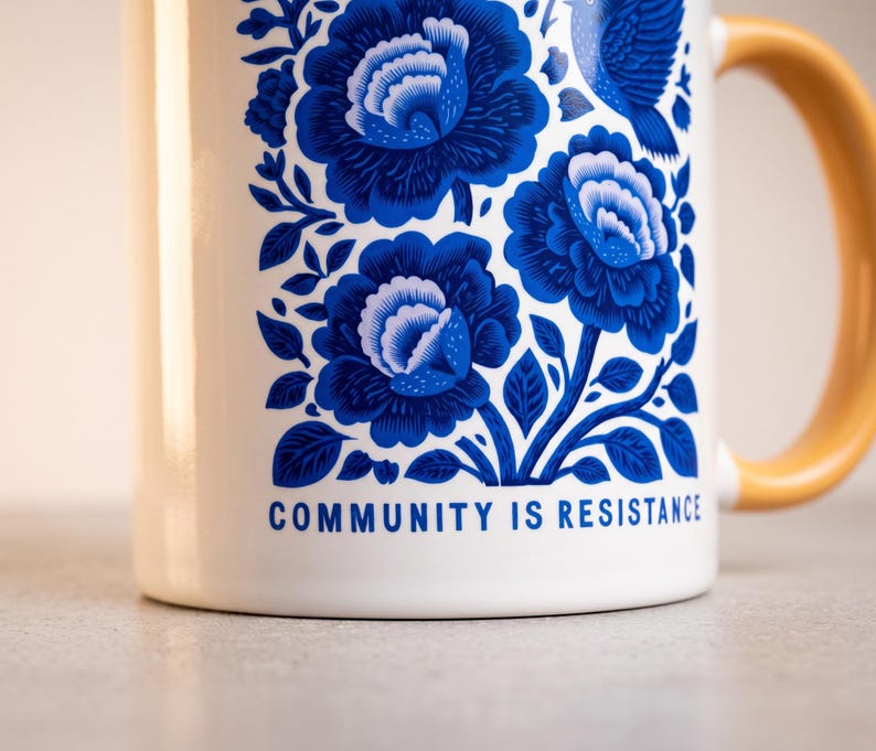 May include: A white ceramic mug with a yellow handle featuring a blue floral pattern of large blossoms and a bird. The text "COMMUNITY IS RESISTANCE" is printed in blue at the bottom of the design. The mug has a smooth, glossy finish throughout.
