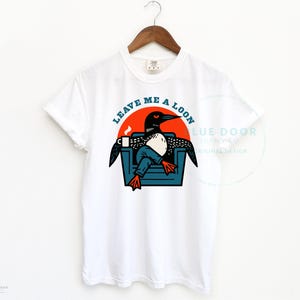 Leave Me A Loon shirt in white hanging on a wooden hanger, funny loon graphic tee for lake lovers and bird watchers, Comfort Colors 1717 unisex fit with retro armchair illustration