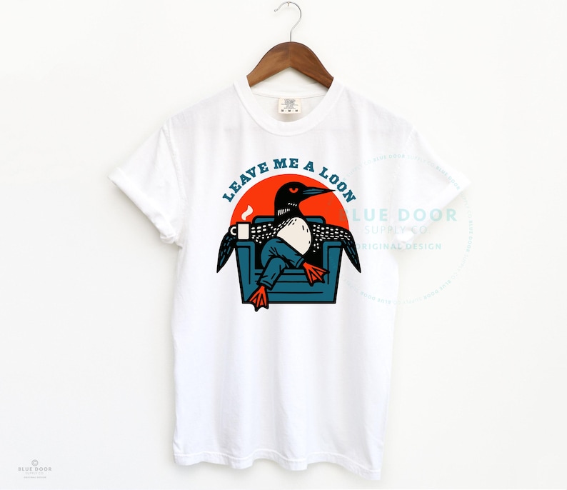 Leave Me A Loon shirt in white hanging on a wooden hanger, funny loon graphic tee for lake lovers and bird watchers, Comfort Colors 1717 unisex fit with retro armchair illustration