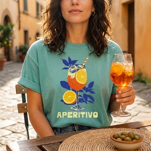 A seafoam comfort colors t-shirt with a graphic of a cocktail with orange slices and the word "APERITIVO" in white. The person is holding a glass of the same cocktail. The background is a sunny outdoor scene.