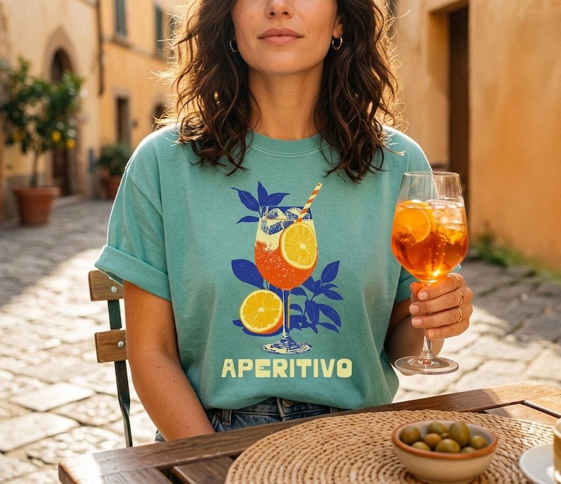 A seafoam comfort colors t-shirt with a graphic of a cocktail with orange slices and the word "APERITIVO" in white. The person is holding a glass of the same cocktail. The background is a sunny outdoor scene.