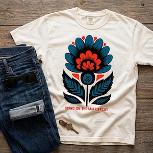 May include: White t-shirt with a large floral design in blue, red, and black, paired with blue denim jeans and sunglasses. The shirt has the text "BRING ON THE MATRIARCHY".