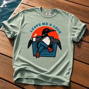 Bird watcher gift shirt in bay green laid across weathered dock boards at an angle, Leave Me A Loon funny graphic tee for birding enthusiasts, retro loon illustration on heavyweight Comfort Colors 1717 cotton