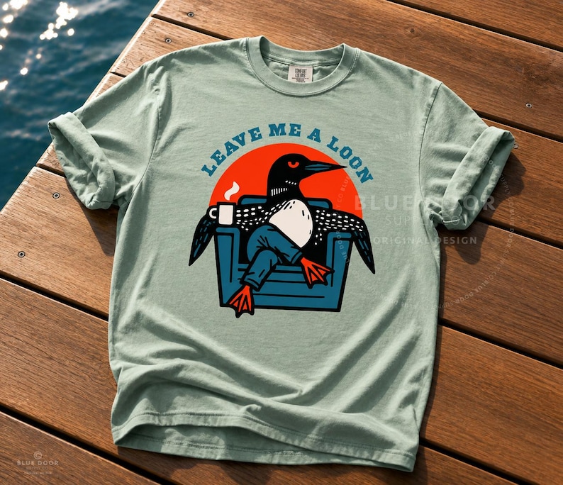 Bird watcher gift shirt in bay green laid across weathered dock boards at an angle, Leave Me A Loon funny graphic tee for birding enthusiasts, retro loon illustration on heavyweight Comfort Colors 1717 cotton