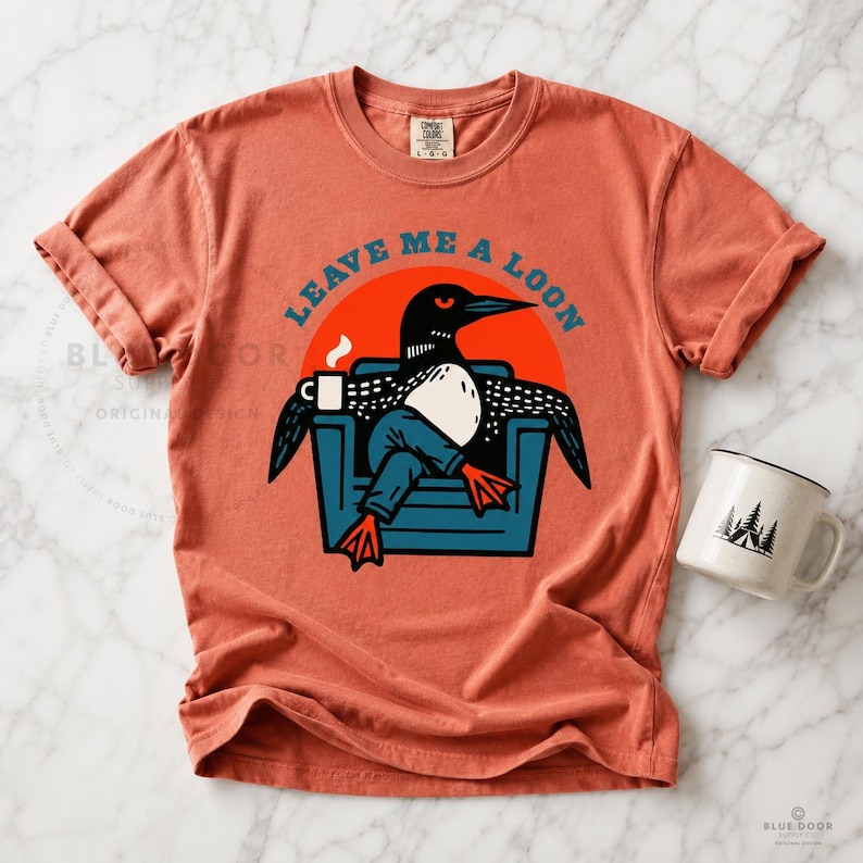 Leave Me A Loon graphic tee in terra cotta orange laid on white marble with a ceramic mug, funny bird lover shirt featuring a loon lounging in an armchair, Comfort Colors 1717 unisex tee for cabin and lake gifts