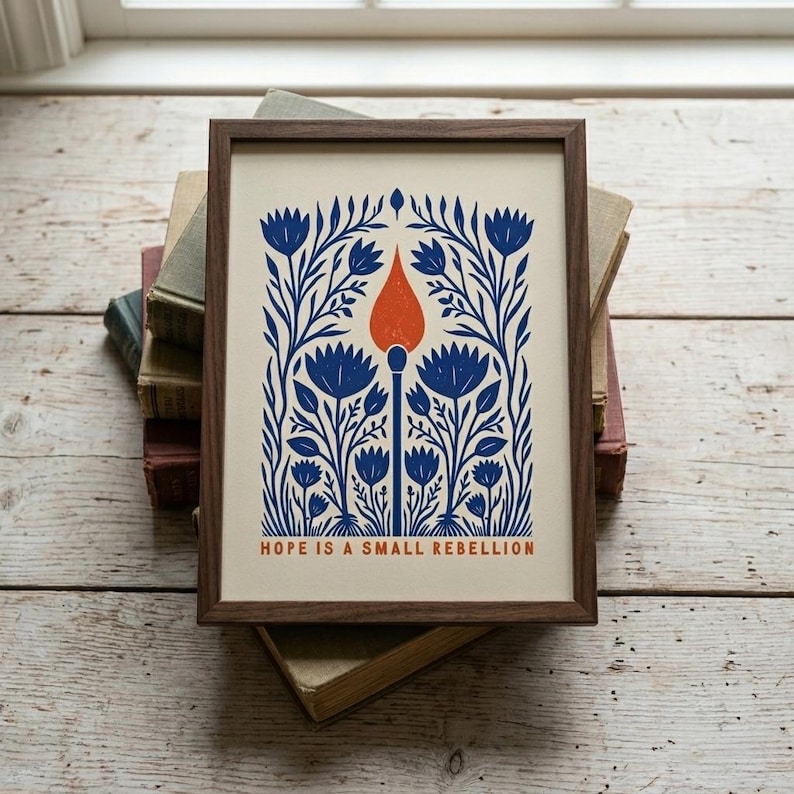 Cobalt blue and orange Hope Is A Small Rebellion art print in a wood frame, a folk-art match-and-flowers illustration. A liberal wall art and civil rights gift print for a reading nook, desk, or bookshelf.