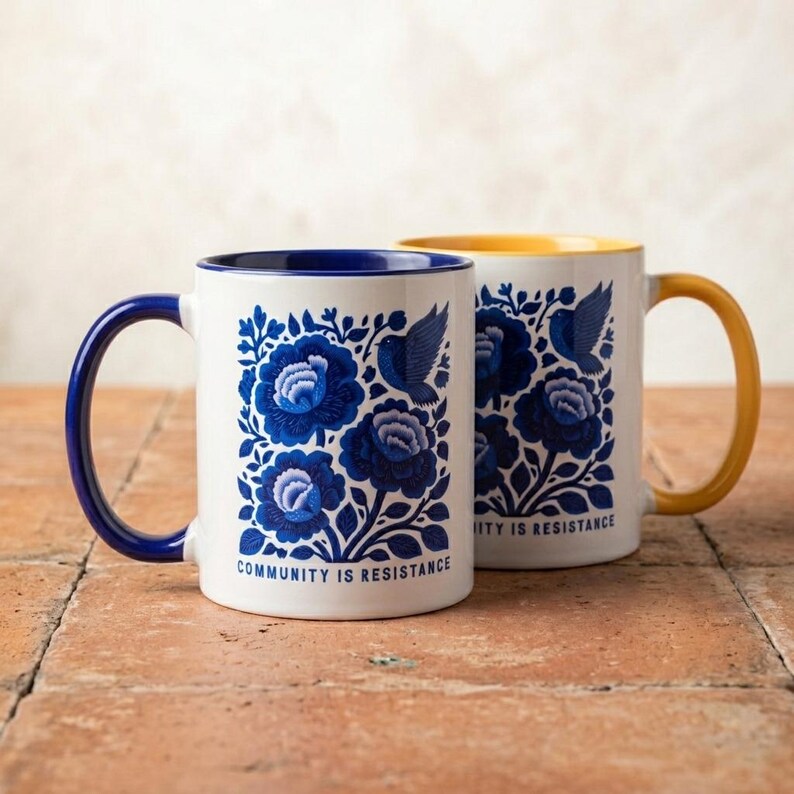 May include: Two white ceramic mugs featuring a blue floral and bird pattern with the text "COMMUNITY IS RESISTANCE" printed at the base. One mug has a dark blue handle and interior, while the other has a yellow handle and interior.