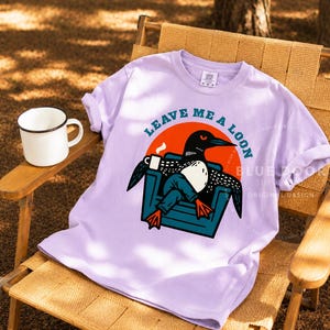Up north gift shirt in orchid draped over a canvas camp chair with a coffee mug on the side table, funny loon graphic tee styled for cabin and lake house living, outdoor lifestyle apparel gift