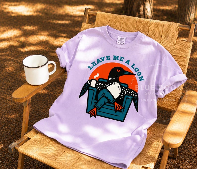 Up north gift shirt in orchid draped over a canvas camp chair with a coffee mug on the side table, funny loon graphic tee styled for cabin and lake house living, outdoor lifestyle apparel gift