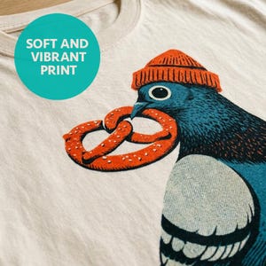 Bird crop top shown up close on ivory, hand-carved block print style linework of a beanie-wearing pigeon with a pretzel