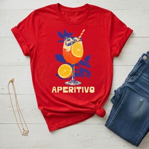 Aperitivo spritz shirt flat lay in red styled with denim and gold necklace — women's Italian summer graphic tee, cocktail lover gift, semi-fitted silhouette