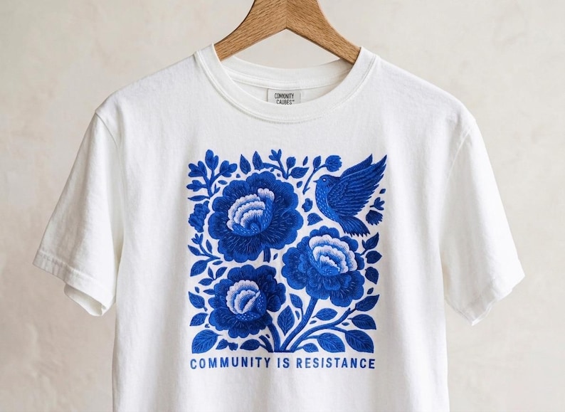 Puede incluir: Una camiseta blanca de manga corta con un estampado gr&aacute;fico azul de tres flores grandes y un p&aacute;jaro. El texto "COMMUNITY IS RESISTANCE" est&aacute; impreso en azul debajo del dise&ntilde;o floral. La camiseta est&aacute; colgada en una percha de madera.
