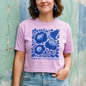 Puede incluir: Una camiseta de manga corta de color morado claro con un gr&aacute;fico cuadrado de flores azul oscuro y un p&aacute;jaro. Debajo del dise&ntilde;o floral, el texto "COMMUNITY IS RESISTANCE" aparece en una fuente negrita de color azul oscuro. La camiseta tiene cuello redondo.