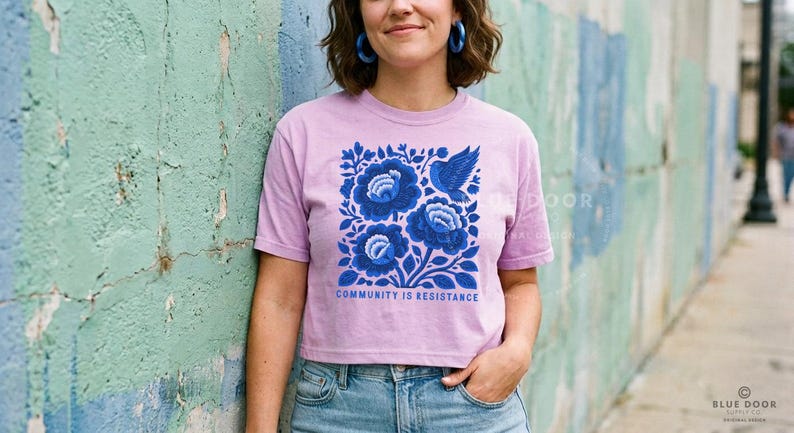 Puede incluir: Una camiseta de manga corta de color morado claro con un gr&aacute;fico cuadrado de flores azul oscuro y un p&aacute;jaro. Debajo del dise&ntilde;o floral, el texto "COMMUNITY IS RESISTANCE" aparece en una fuente negrita de color azul oscuro. La camiseta tiene cuello redondo.