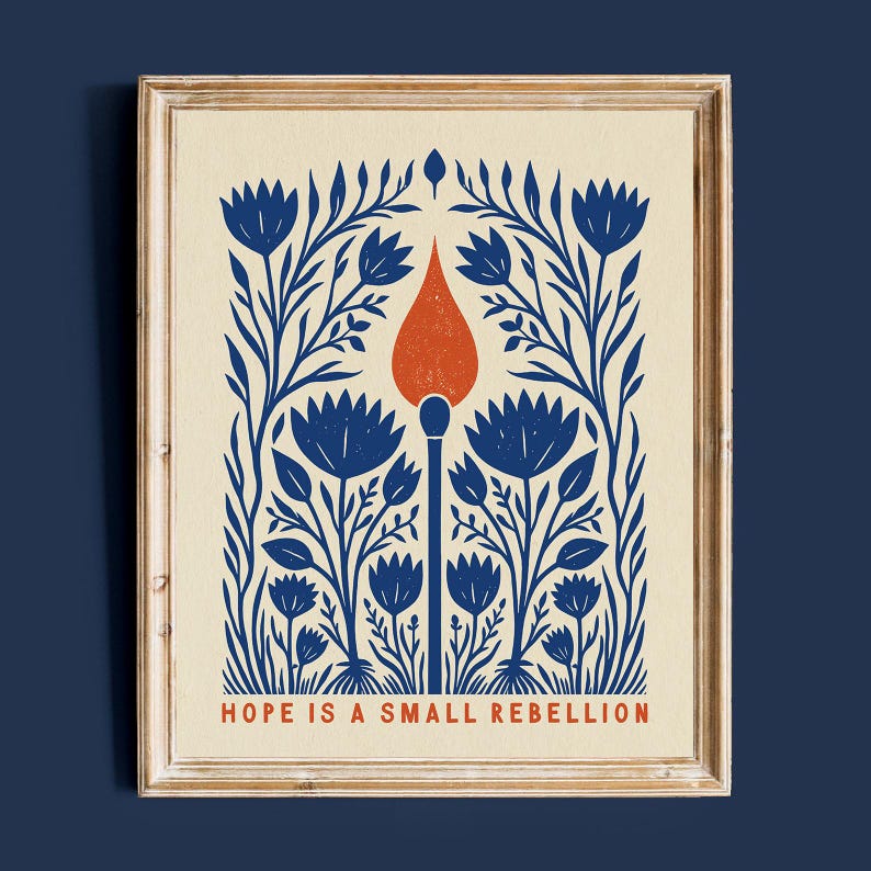 Hope Is A Small Rebellion folk art print framed in light wood on a deep navy wall. Blue and orange wildflower-and-match illustration, a social justice and feminist art print for a gallery wall or activist's home.