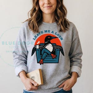Woman wearing a sport gray loon sweatshirt while holding a book, cozy camping sweatshirt with funny loon illustration for lake house and cabin gifting