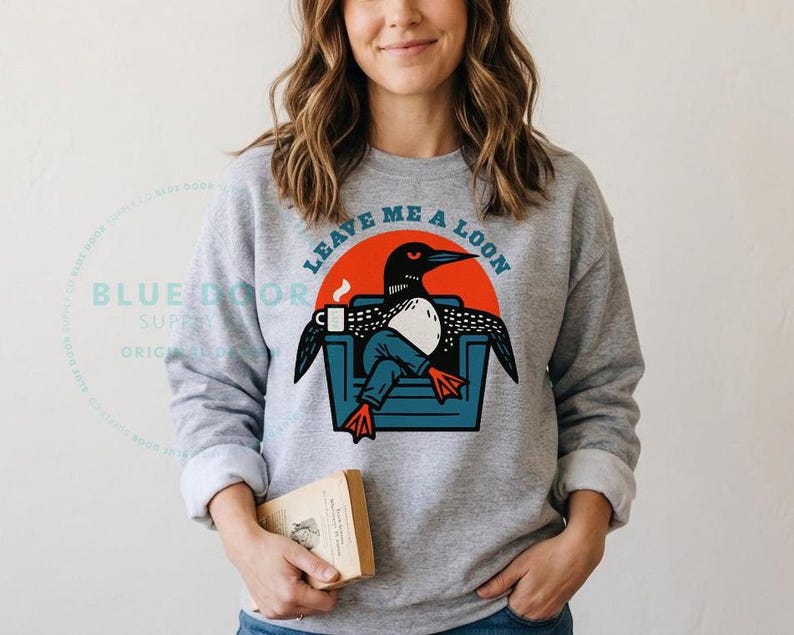 Woman wearing a sport gray loon sweatshirt while holding a book, cozy camping sweatshirt with funny loon illustration for lake house and cabin gifting