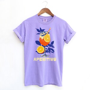 A lavendar purple t-shirt featuring a graphic of an Aperitivo cocktail with orange slices and a straw. The word "APERITIVO" is printed in yellow below the cocktail illustration. The shirt is hanging on a wooden hanger.