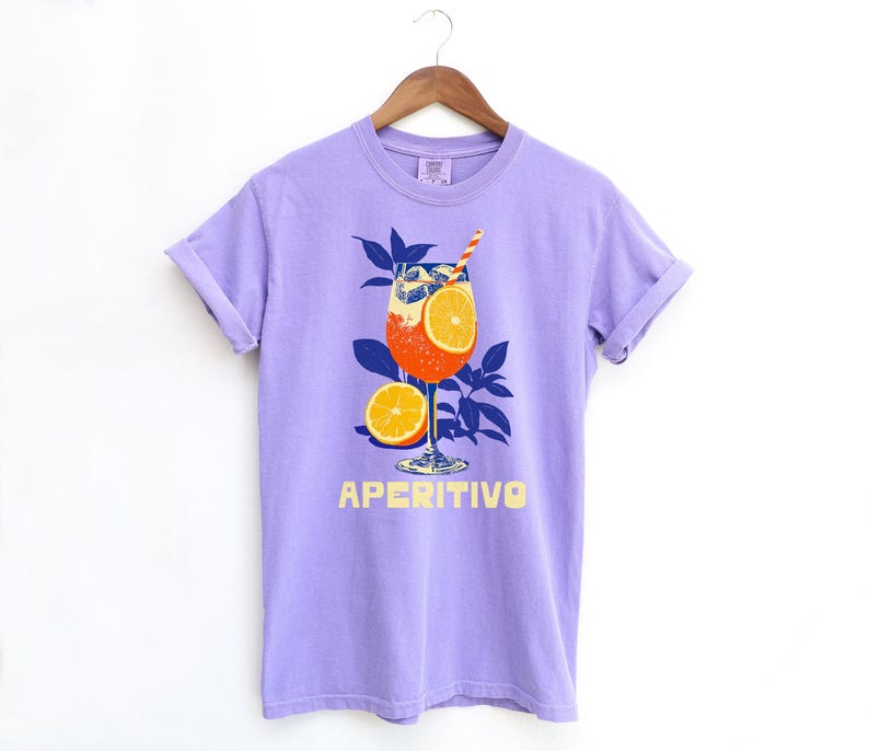 A lavendar purple t-shirt featuring a graphic of an Aperitivo cocktail with orange slices and a straw. The word "APERITIVO" is printed in yellow below the cocktail illustration. The shirt is hanging on a wooden hanger.