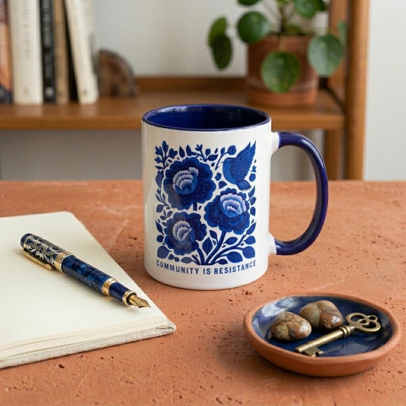May include: A white ceramic mug with a dark blue interior and handle, featuring a blue floral pattern with a bird and the text "COMMUNITY IS RESISTANCE" printed in blue. The mug sits alongside a blue and gold fountain pen and a small blue dish.