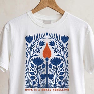 White Comfort Colors boxy crop tee on a wooden hanger featuring a bold folk art botanical illustration of a lit match with red-orange flame surrounded by wildflowers, reading Hope Is A Small Rebellion — feminist liberal graphic crop top.
