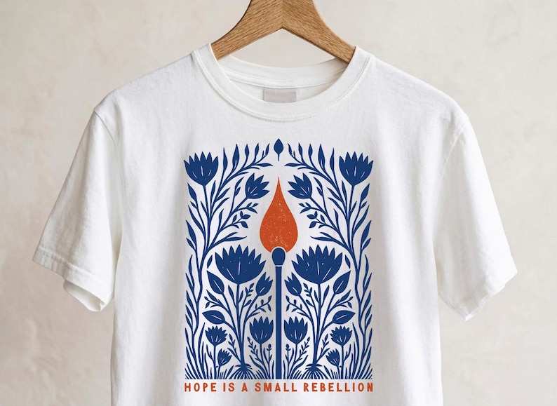 White Comfort Colors boxy crop tee on a wooden hanger featuring a bold folk art botanical illustration of a lit match with red-orange flame surrounded by wildflowers, reading Hope Is A Small Rebellion — feminist liberal graphic crop top.
