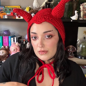 May include: A handmade red crochet hat with devil horns. The hat features a scalloped edge and ties under the chin. The hat is a unique accessory.