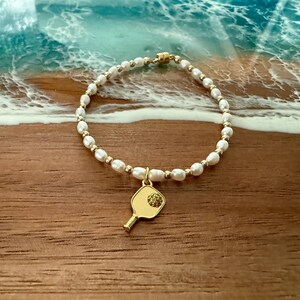 May include: A pearl bracelet with gold accents and a gold pickleball paddle charm. The bracelet features white pearls and small gold beads. The paddle charm has a textured surface and a small, round embellishment.