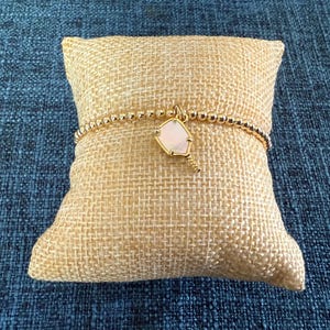 May include: A gold beaded bracelet with a pendant featuring a light pink, shield-shaped stone. The bracelet is displayed on a small, square, tan burlap pillow against a blue textured background.