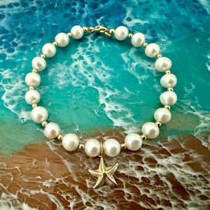 May include: A circular bracelet featuring white spherical beads alternating with small gold-toned metal spacers. A gold-toned starfish charm hangs from the bottom of the bracelet, which is secured with a gold-toned metal lobster claw clasp.