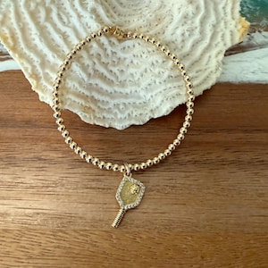 May include: Gold beaded bracelet with a charm shaped like a pickleball paddle. The paddle charm is embellished with small, clear stones. The bracelet is displayed on a wooden surface with a white, textured object.