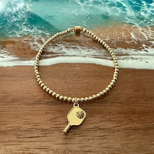 May include: Gold beaded bracelet with a pickleball paddle charm. The charm is a gold-colored pickleball paddle with a textured center. The bracelet is on a wooden surface with a blurred ocean background.